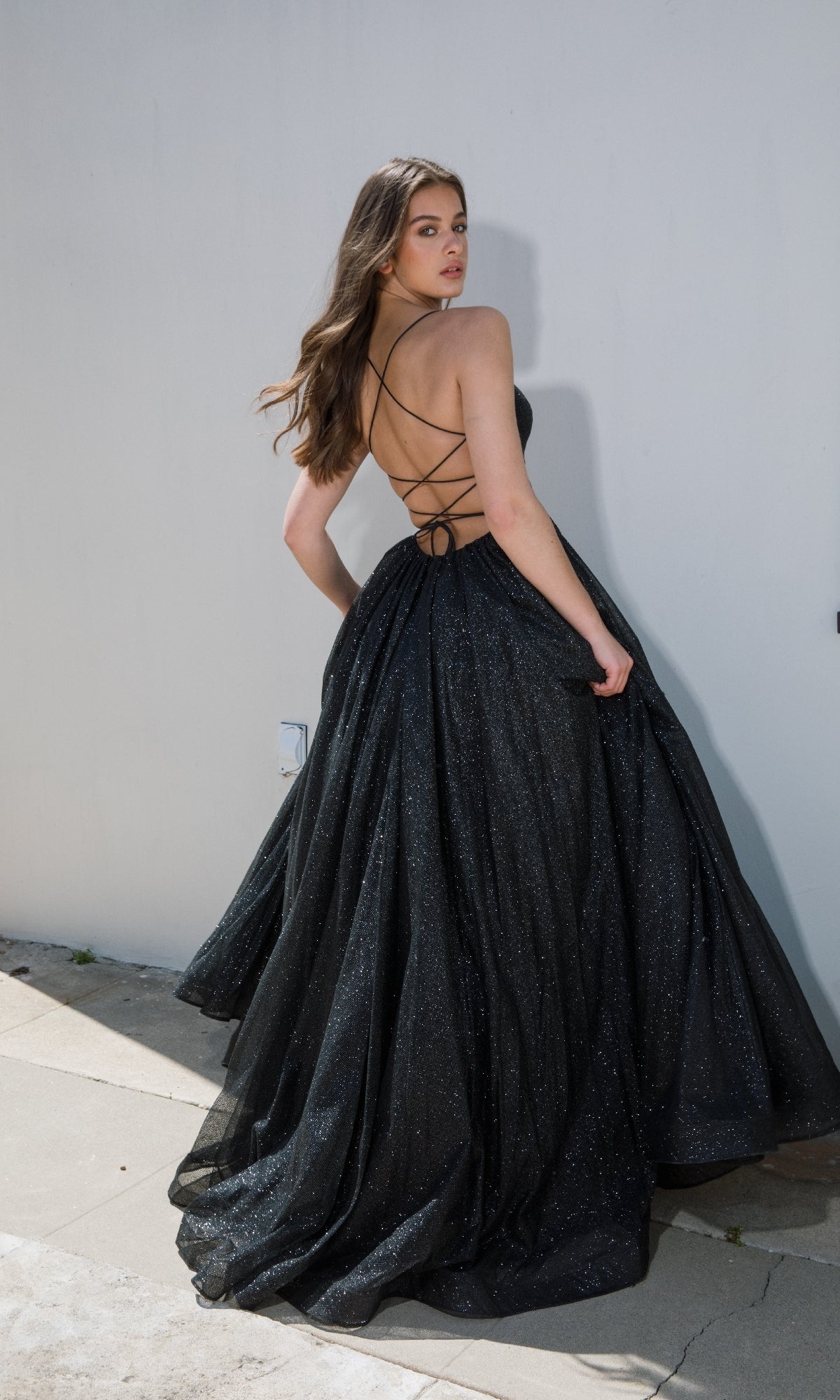 Model wears the Strappy-Back Glitter Prom Ball Gown Eureka 9009, posing outdoors against a white wall and looking over her shoulder, showcasing its black glitter fabric and lace-up open back.